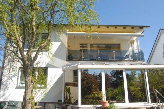 Balkon der Ferienwohnung in der 1.Etage Helles modernes Haus mit Balkon und großer Fensterfront, umgeben von Bäumen.