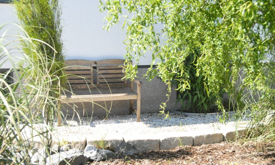 Vorgarten Holzbank umgeben von Pflanzen und Kies, vor einer hellen Wand.