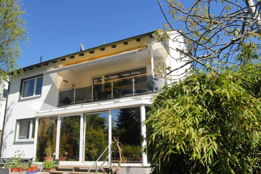 Gartenansicht Moderne Villa mit Balkon, umgeben von Bäumen und grünem Garten.